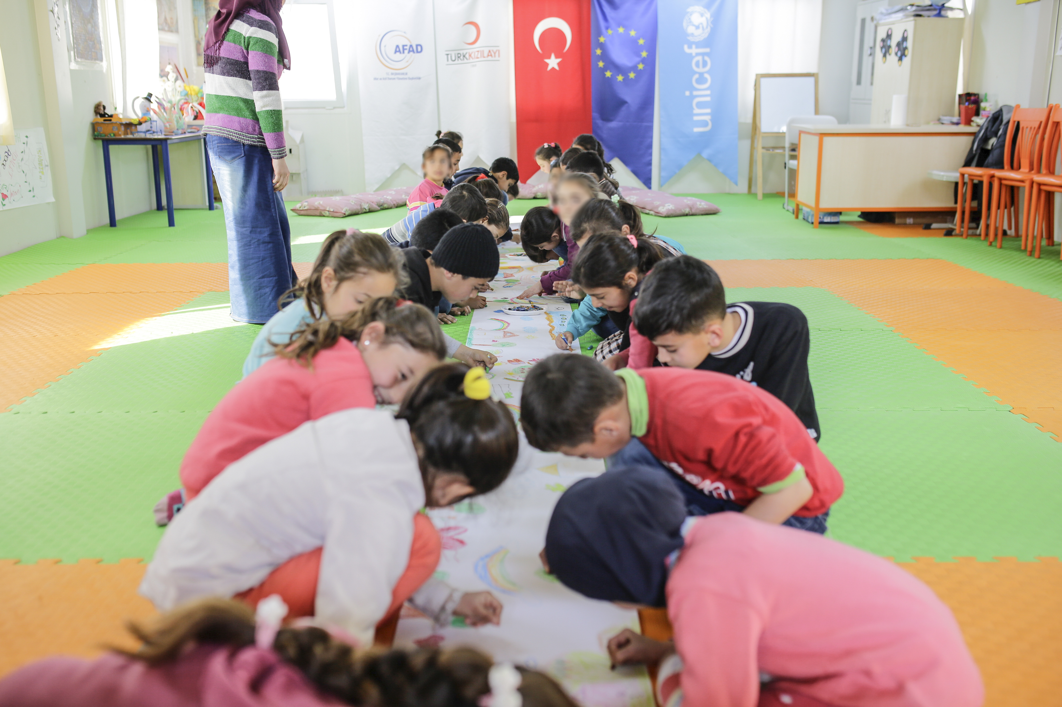 Syyrialaislapsia piirtämässä. Taustalla Turkin, EU:n ja Unicefin tunnukset.