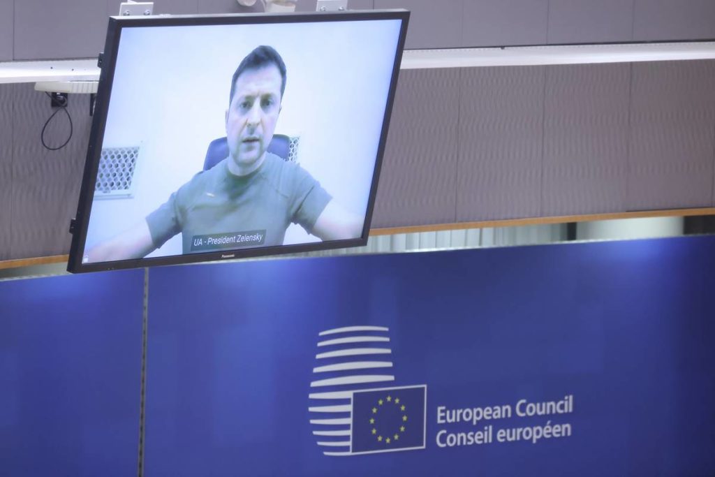 Kuvassa on tietokoneen näyttö seinällä. Siinä näkyvät Ukrainan presidentin Volodymyr Zelenskyin kasvot. Taustalla Eurooppa-neuvoston logoseinä.